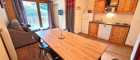 Un espace de vie compact comprenant une table à manger, des chaises, un canapé, un coin cuisine avec des meubles en bois et des appareils électroménagers, ainsi qu’une porte-fenêtre coulissante donnant sur un espace extérieur.