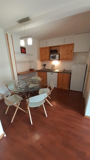 Cozinha compacta com armários de madeira, micro-ondas, frigorífico e mesa de jantar de vidro com cadeiras.