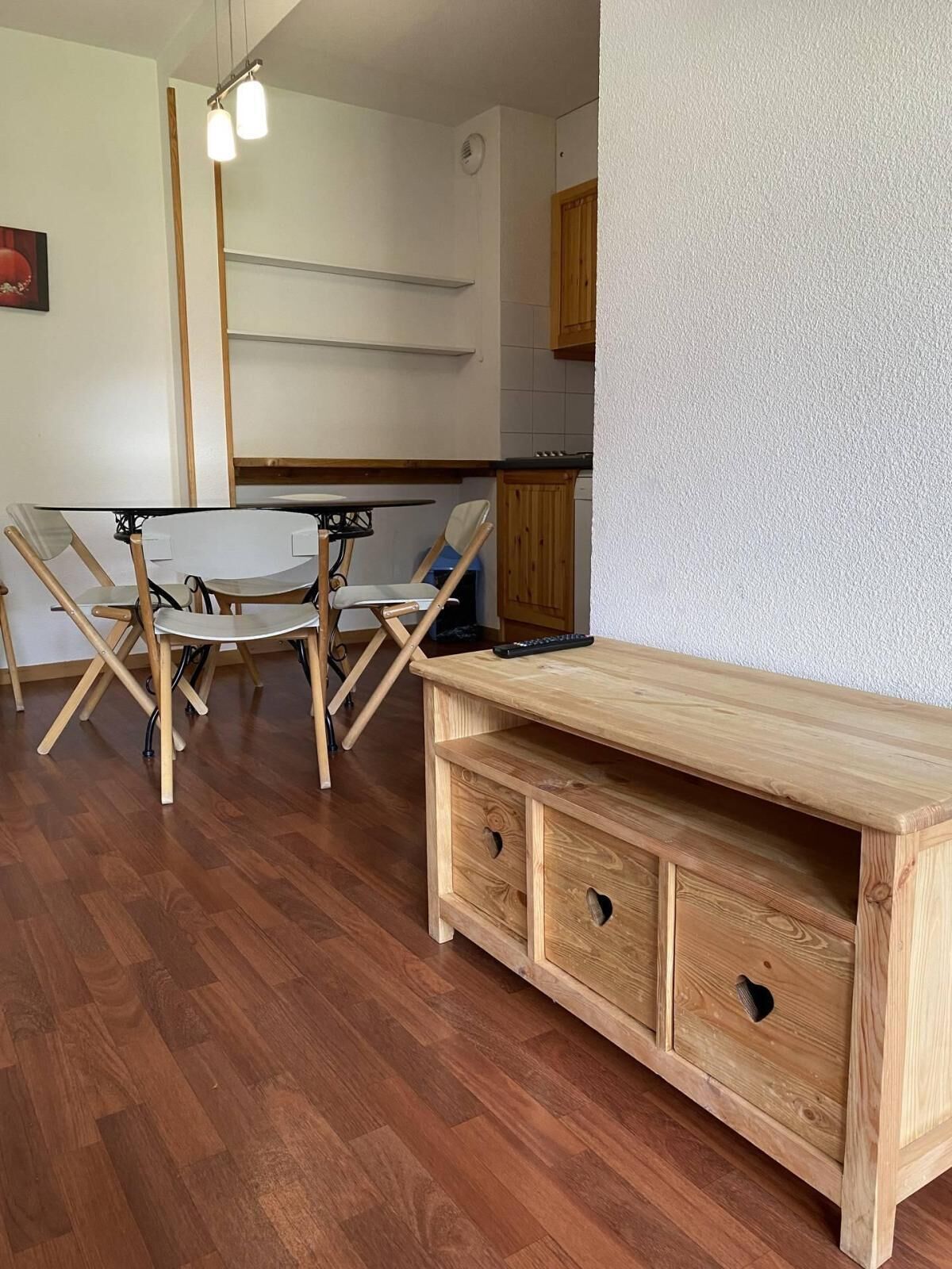 A dining area with a glass table and wooden chairs, a wooden cabinet with drawers, and a kitchen in the background.