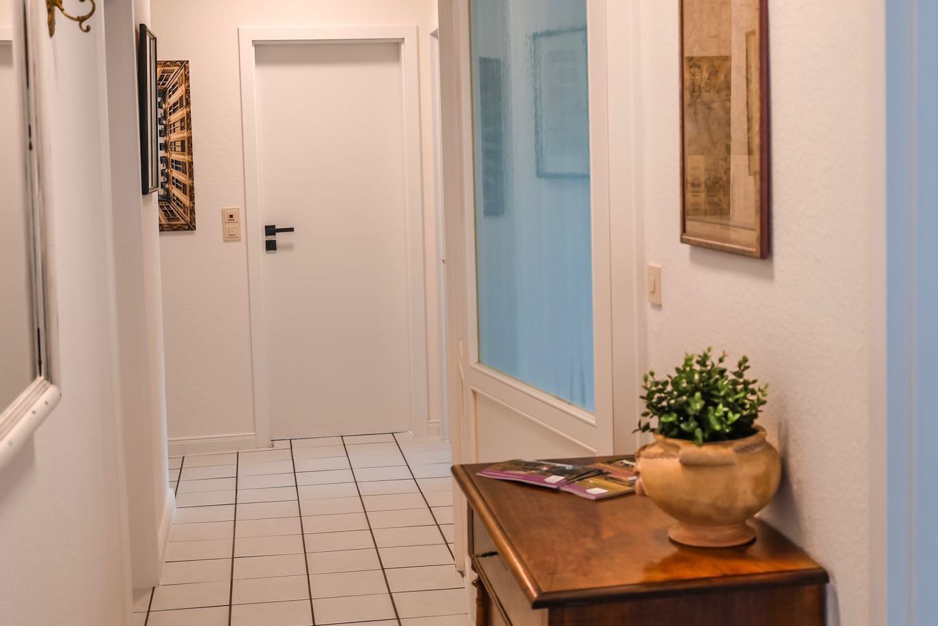 Um corredor com uma porta branca, uma janela com cortinas azuis e uma mesa de madeira com uma planta e alguns objetos.