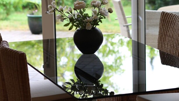 Una mesa de vidrio con un jarrón de flores blancas, sillas de mimbre y vistas al verdor exterior.