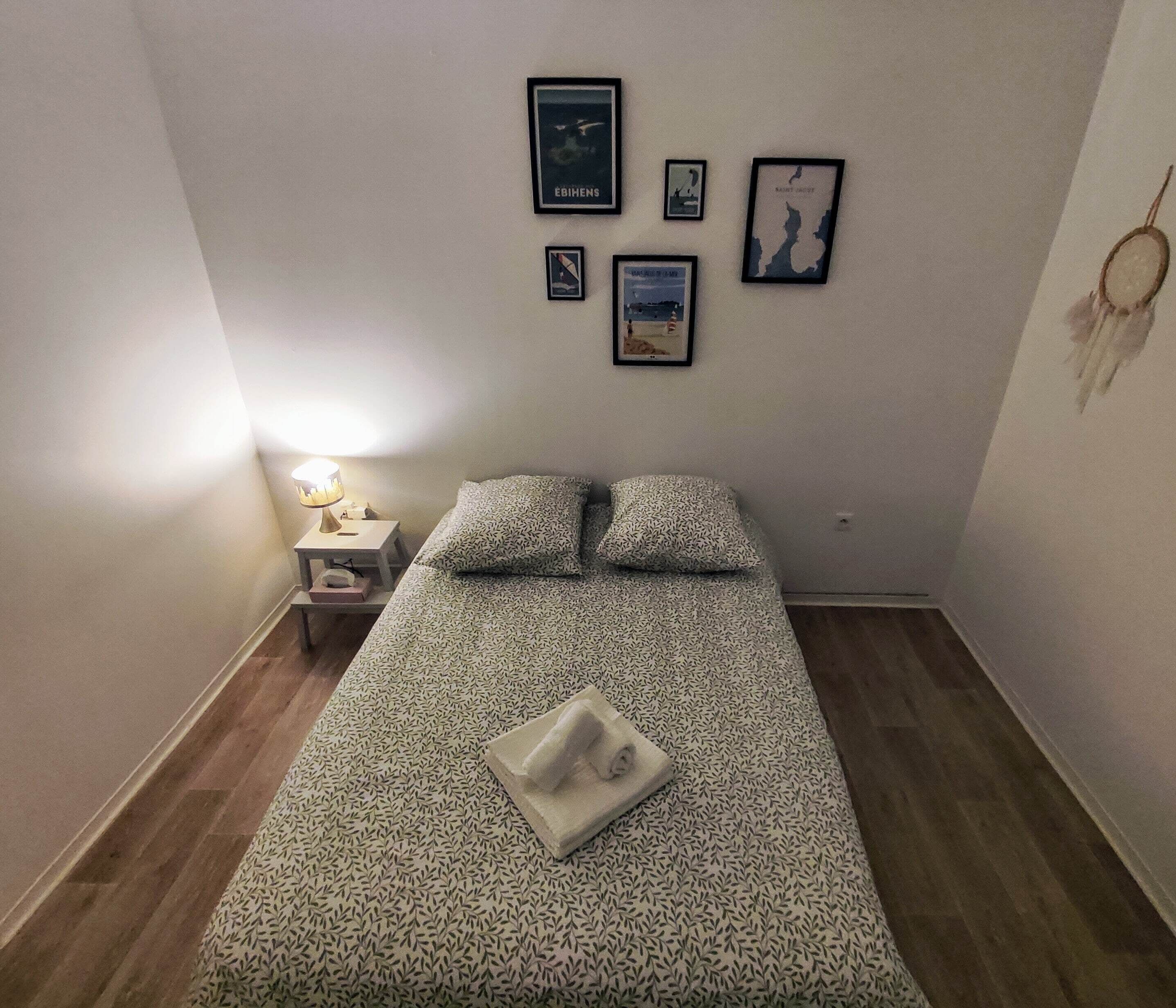 A small, neatly arranged bedroom with a bed, a nightstand, a lamp, and several framed pictures on the wall.