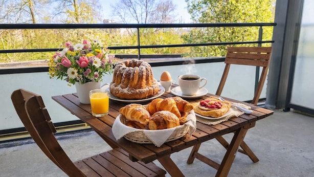 A wooden table with breakfast items including pastries, a cake, and beverages, set on a balcony with a view of trees. - Le Salluste: T2 cosy with balcony in Strasbourg. (Strasbourg)
