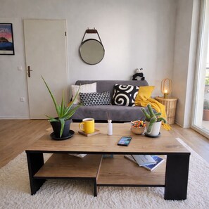 A living room with a grey sofa, a round mirror, a coffee table with a yellow mug, and various plants.