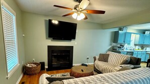 A living room with a sofa, ottoman, fireplace, ceiling fan, and a kitchen area in the background.