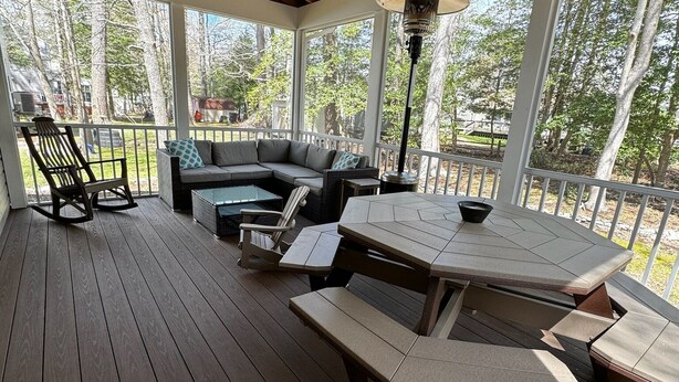 A covered outdoor deck with a wooden floor, a sectional sofa, a dining table with benches, and a ceiling fan.