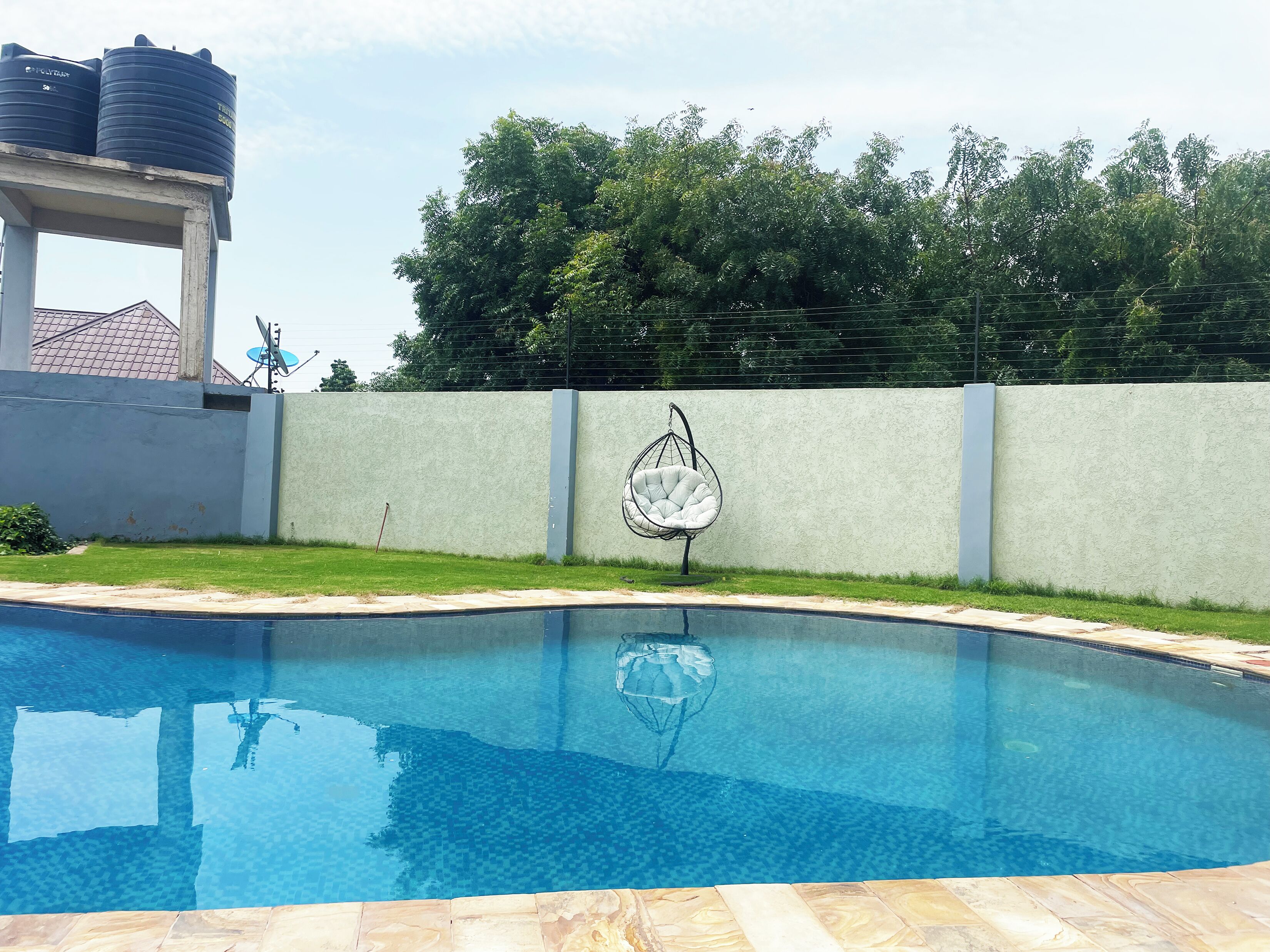 A swimming pool with a hanging chair, a water tank, and a wall with trees in the background.