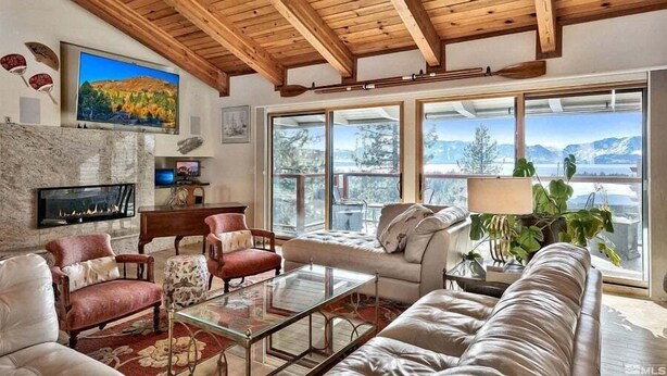 A spacious living room with a fireplace, comfortable seating, large windows offering mountain views, and a wooden ceiling. - Lx121 Pristine Tahoe Experience (Zephyr Cove)