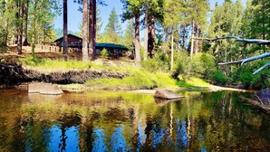A cabin with a deck overlooking a clear stream, surrounded by tall trees and a rocky shoreline.