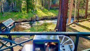 A Weber Spirit grill on a wooden deck with a view of a forest and a stream.