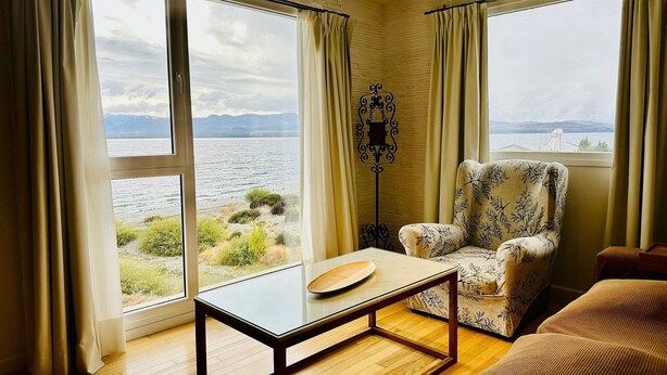 Una habitación con ventanas amplias, suelo de madera, un sillón estampado, una mesa y una lámpara de pie.