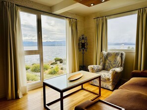 A room with large windows, a wooden floor, a patterned armchair, a table, and a floor lamp.
