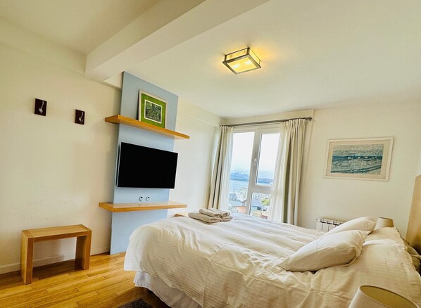 A bedroom with a bed, a TV mounted on a wall, a window with curtains, and a view of the sea.