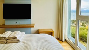 Un dormitorio con una cama, un televisor montado en la pared, una mesita pequeña y un ventanal con vistas al mar.
