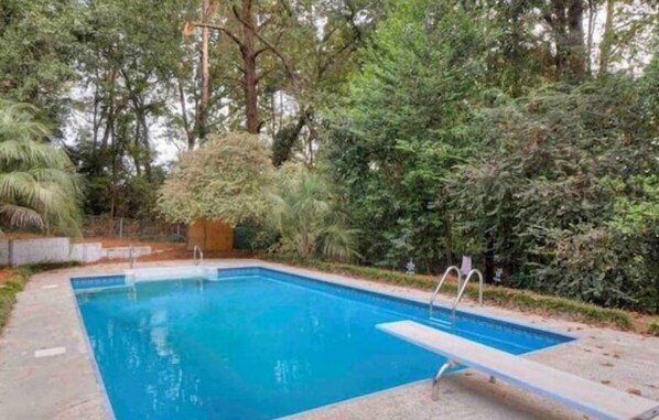 A rectangular swimming pool with a clear blue water surface, surrounded by a concrete deck and bordered by dense greenery and trees. - Charming 3BR Home + Private Pool (North Augusta)