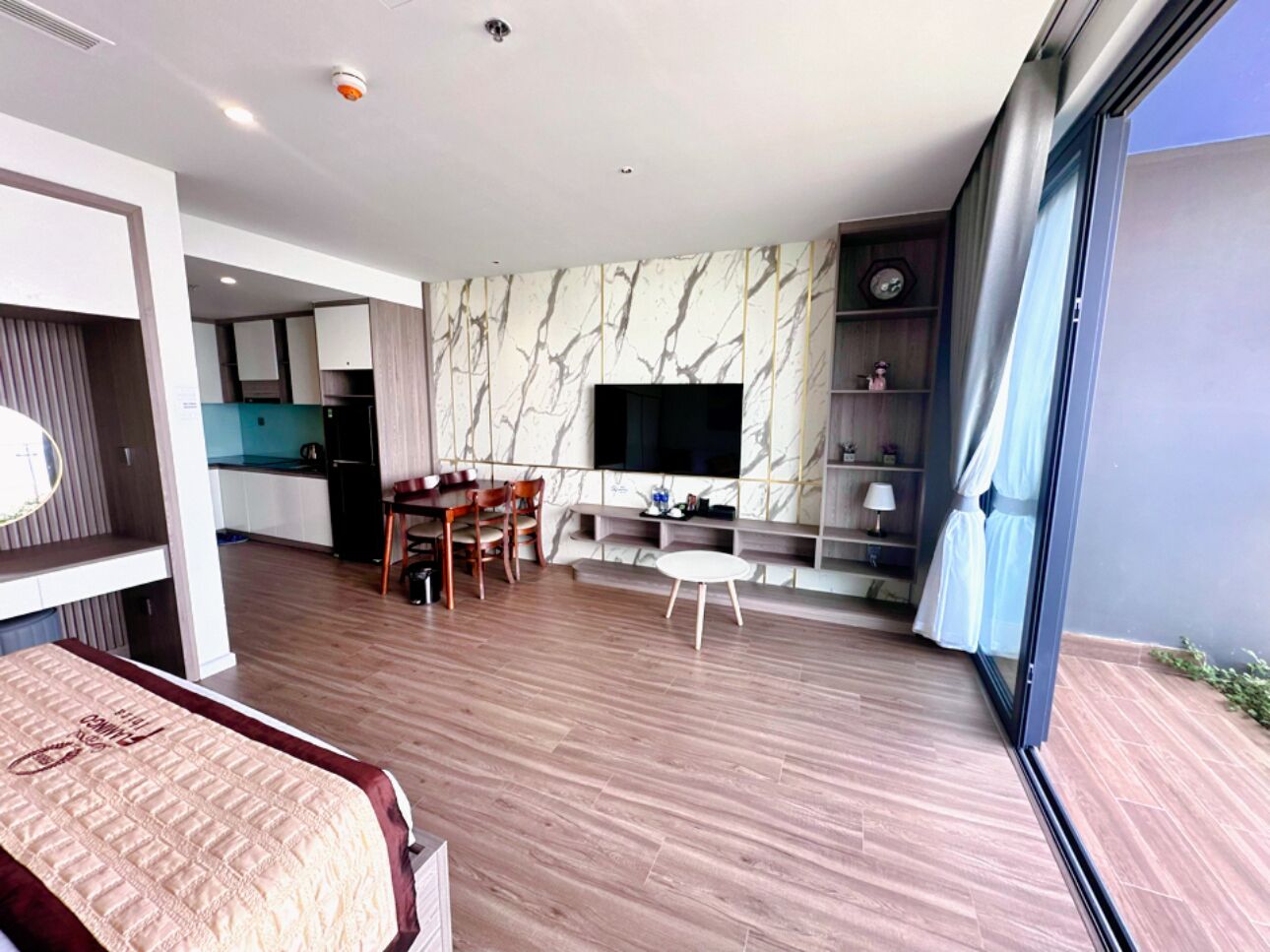 A modern hotel room with a large bed, a dining area with a table and chairs, a TV mounted on the wall, and a balcony with a view.