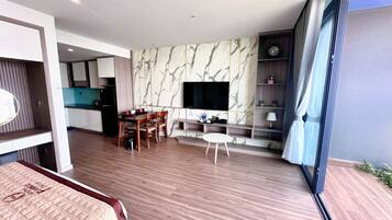 A modern hotel room with a large bed, a dining area with a table and chairs, a TV mounted on the wall, and a balcony with a view.