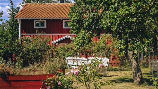 Una casa roja con techo de tejas, rodeada de un jardín con bancos blancos y flores rosadas.