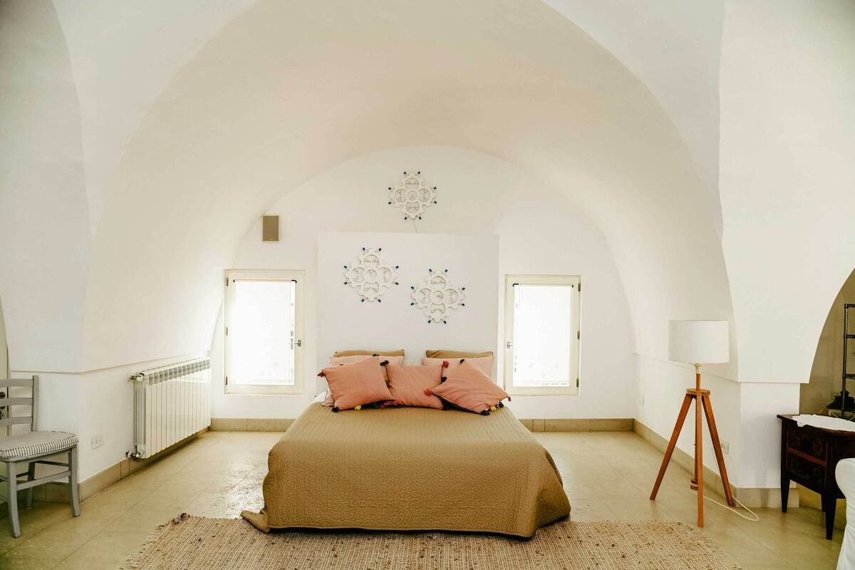 A bedroom with a large bed, two windows, a radiator, a chair, a lamp, and a decorative wall piece.