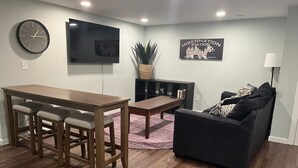 Un salon moderne avec une table à manger en bois, des tabourets de bar, un canapé noir, un téléviseur fixé au mur et une plante en pot.