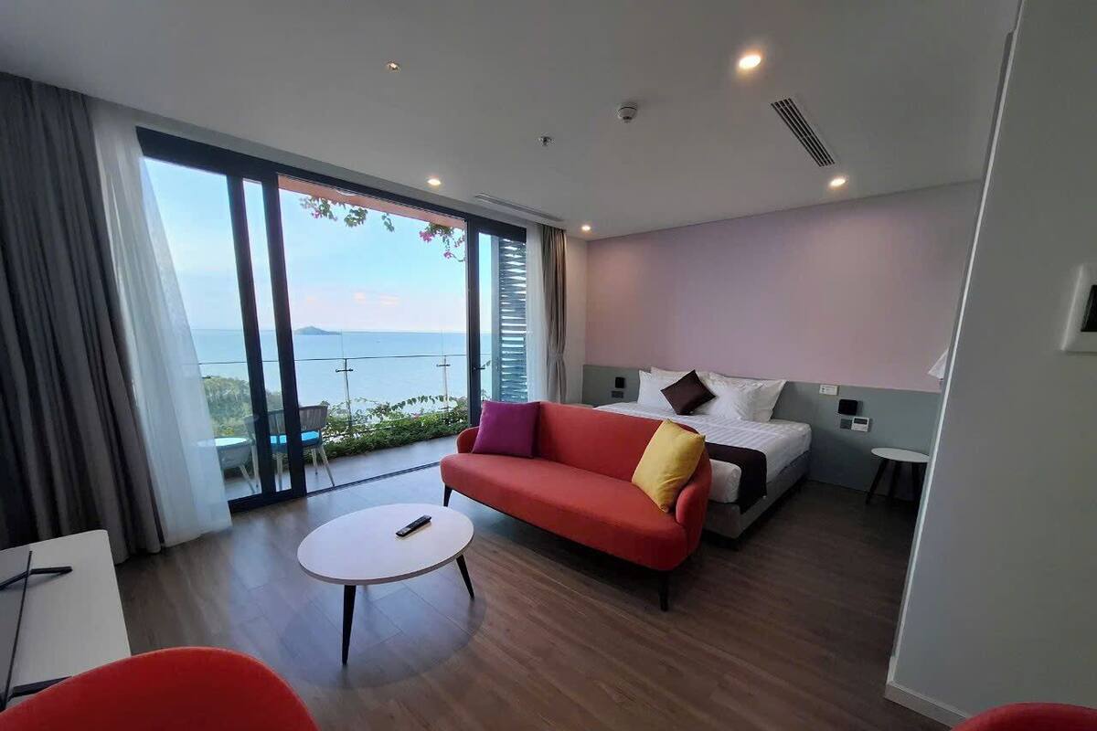 A modern hotel room with a large sliding glass door leading to a balcony, a red sofa, a small round table, and a bed with a white headboard.