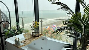 Un baño con un amplio ventanal con vista a la playa, una bañera y una bandeja de madera con artículos de aseo.