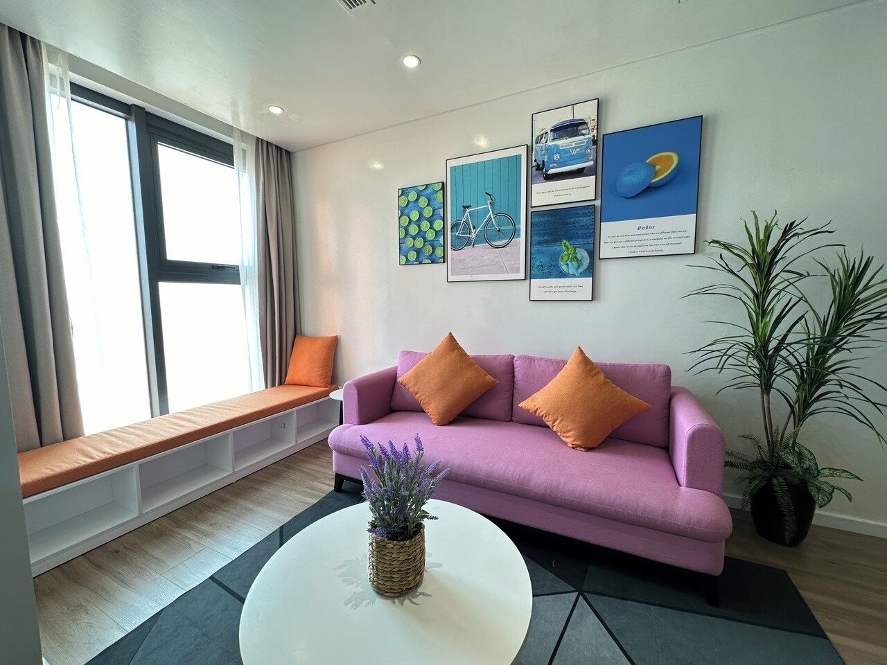 Una sala de estar moderna con un sofá púrpura, cojines naranjas, una mesa de centro blanca y un banco junto a la ventana.