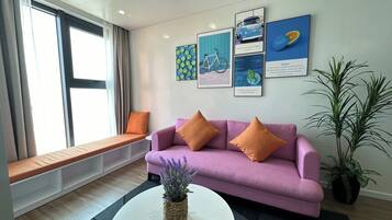 Una sala de estar moderna con un sofá púrpura, cojines naranjas, una mesa de centro blanca y un banco junto a la ventana.