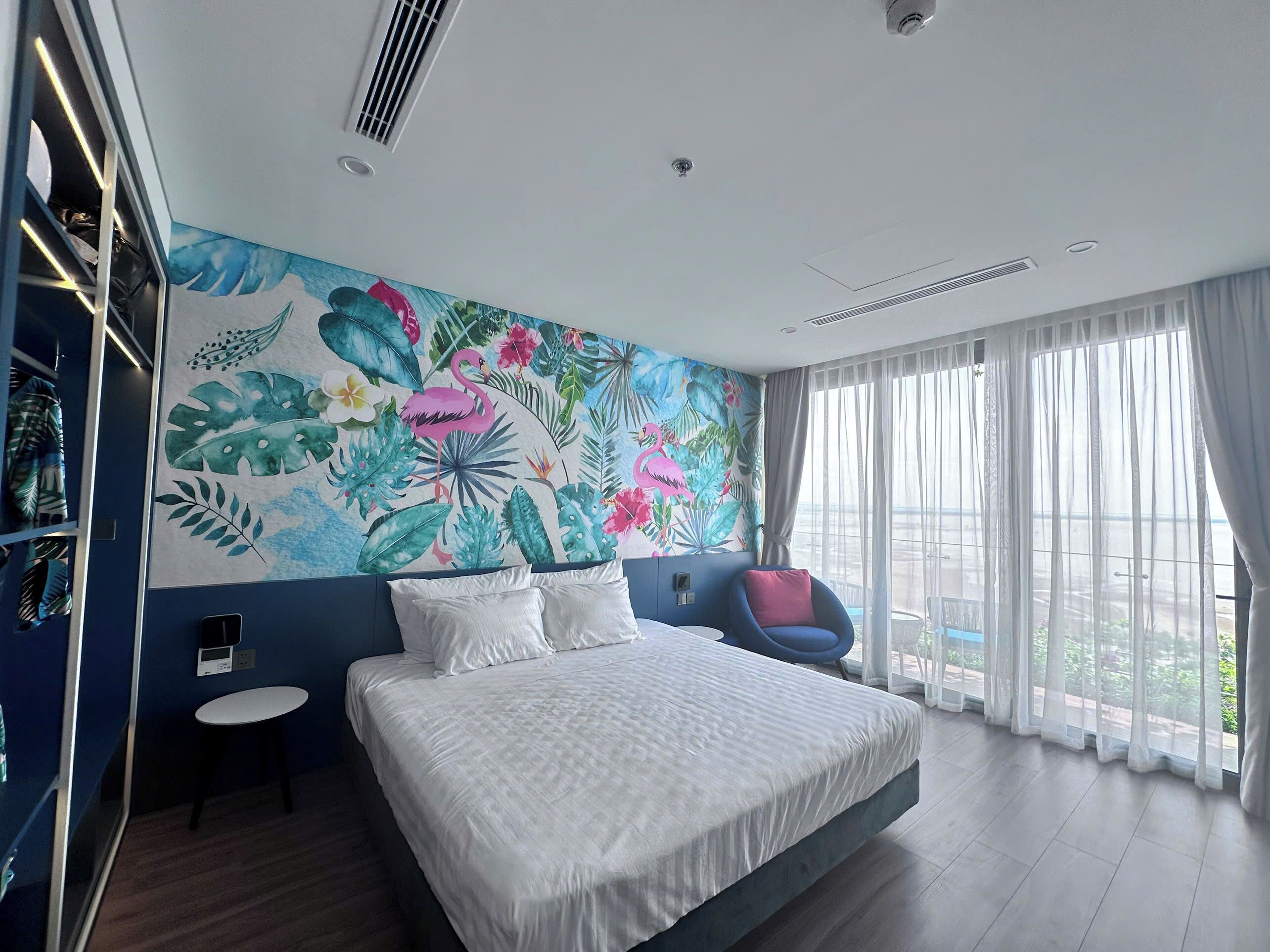 Un dormitorio moderno con una cama grande, un mural tropical, una mesita y vistas al exterior a través de cortinas translúcidas.