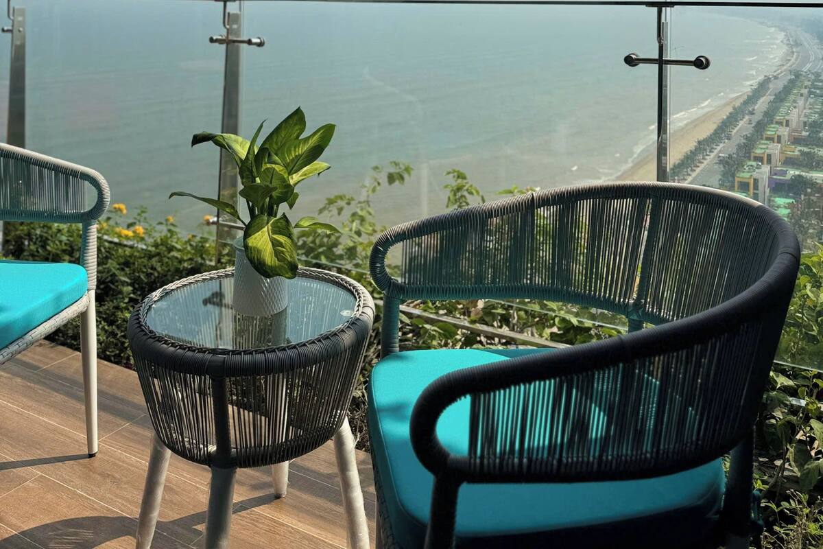 A balcony with wicker chairs and a glass table, overlooking a beach and cityscape.