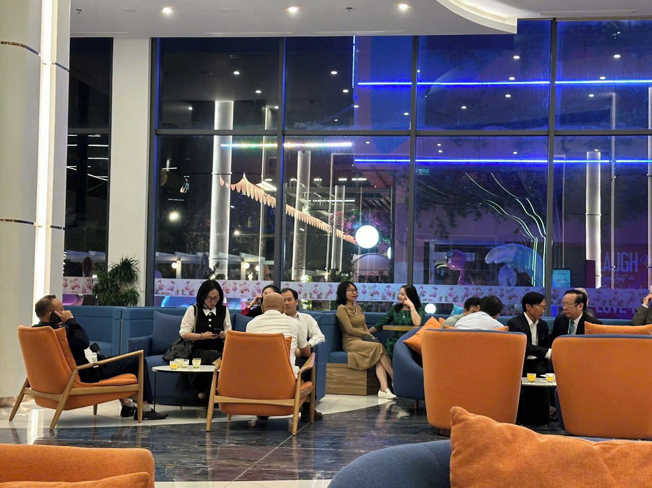 A modern indoor seating area with orange and blue furniture, large windows, and a decorative ceiling light fixture.