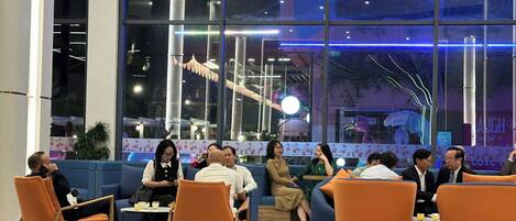 A modern indoor seating area with orange and blue furniture, large windows, and a decorative ceiling light fixture.