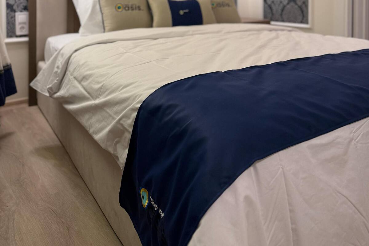 A hotel room with a large bed, a headboard with a logo, patterned wallpaper, and wall-mounted lamps.