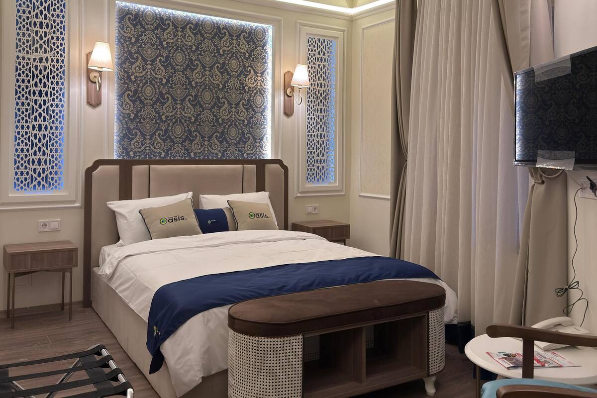 A hotel room with a bed, bedside tables, a desk, a chair, a TV, and a patterned rug.