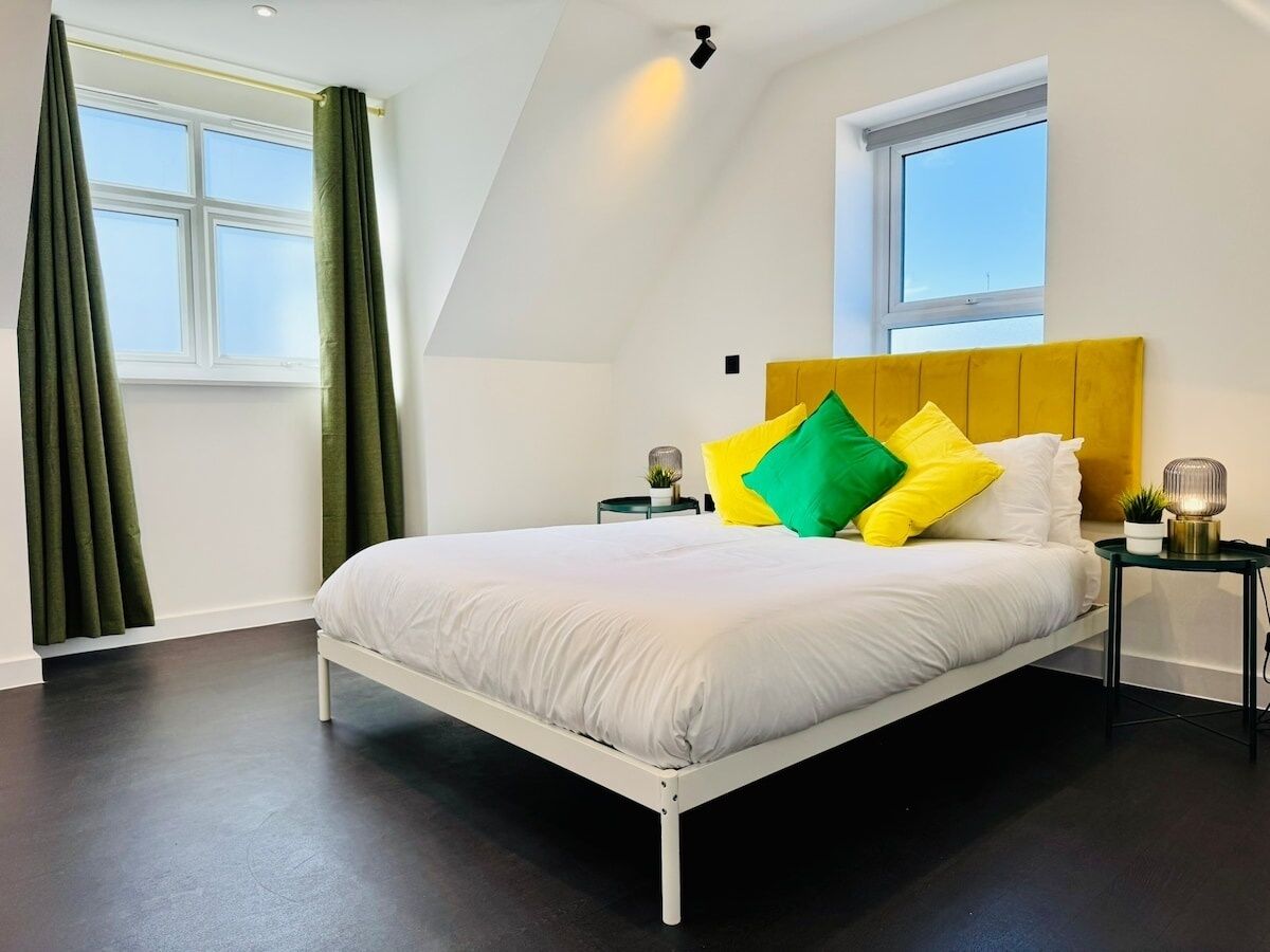 A modern bedroom with a white bed, green curtains, a yellow headboard, and a small table with a lamp.
