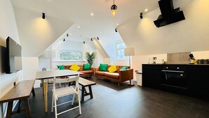 A modern loft apartment with a dining area, sofa set, kitchen, and a large window providing natural light.