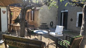 Zona d'estar a l'aire lliure de disseny rústic, amb una cadira de fusta, taula i caires, tot envoltat per un mur de pedra i un arbre.