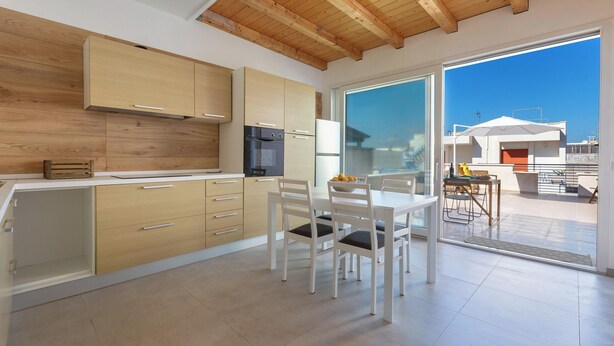 Cocina moderna con armarios de madera, comedor con sillas blancas y un amplio ventanal corredero que da a un patio exterior.