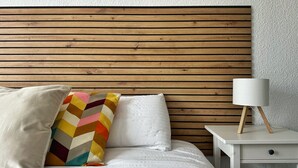 A bed with a wooden headboard, a white pillow, a colorful pillow, and a bedside table with a lamp. - Tierruca Green 30023 (Somo)