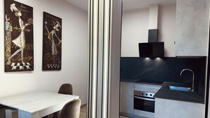 A modern kitchen with a black countertop, stainless steel appliances, and a white dining area with a table and chairs. - Flower Deluxe - Garden Apartment-6082B (Noja)