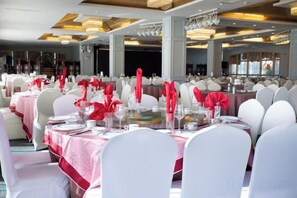 Un salón de banquetes con mesas redondas dispuestas para comer, sillas blancas y servilletas rojas.