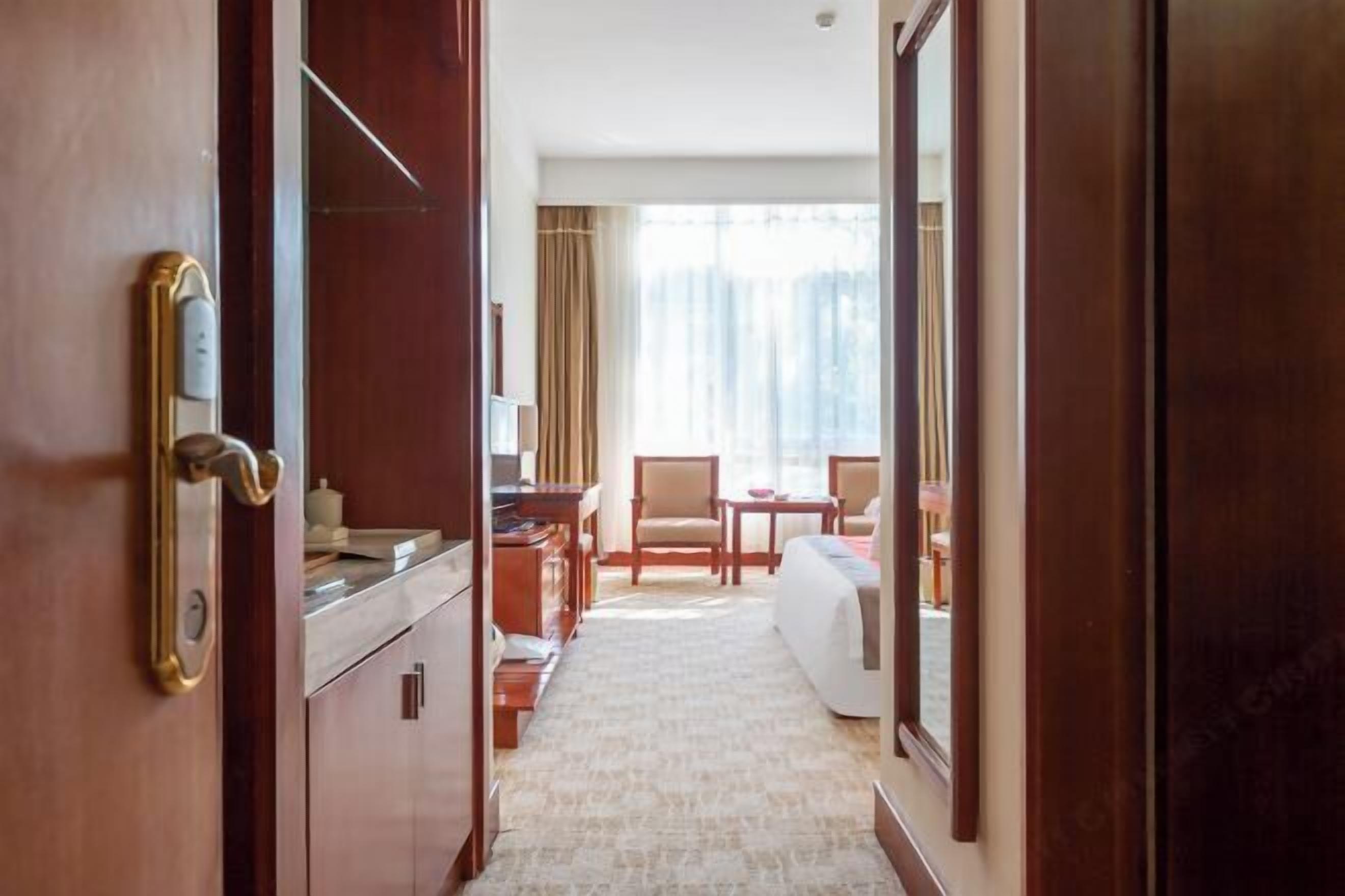 Quarto de hotel com porta de madeira, uma escrivaninha, uma cadeira, uma cama e vista exterior por grandes janelas.