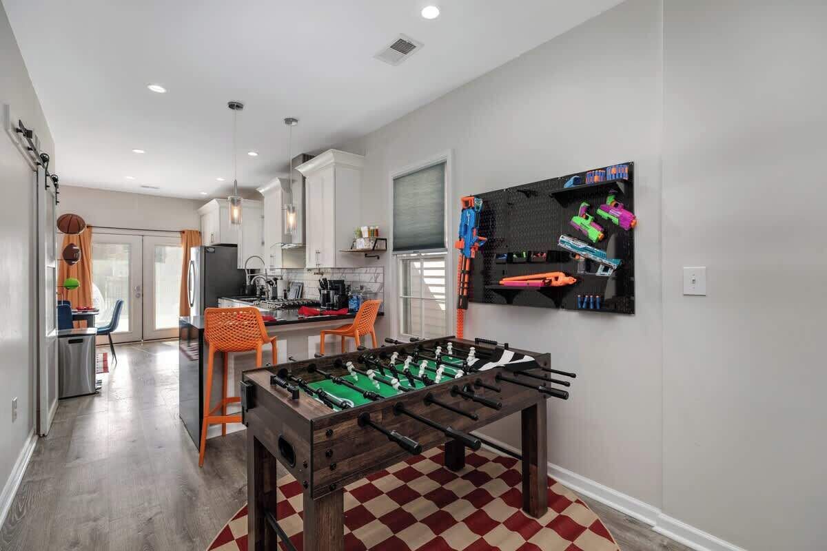 A modern kitchen with a foosball table, a dining area with orange chairs, and a wall-mounted toy shelf.