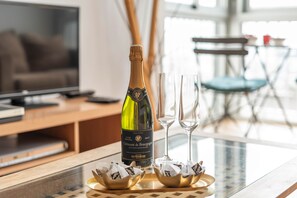Uma garrafa de Crémant de Bourgogne, dois copos de vinho vazios e uma taça de chocolates sobre uma mesa num quarto com televisão e vista para o exterior.