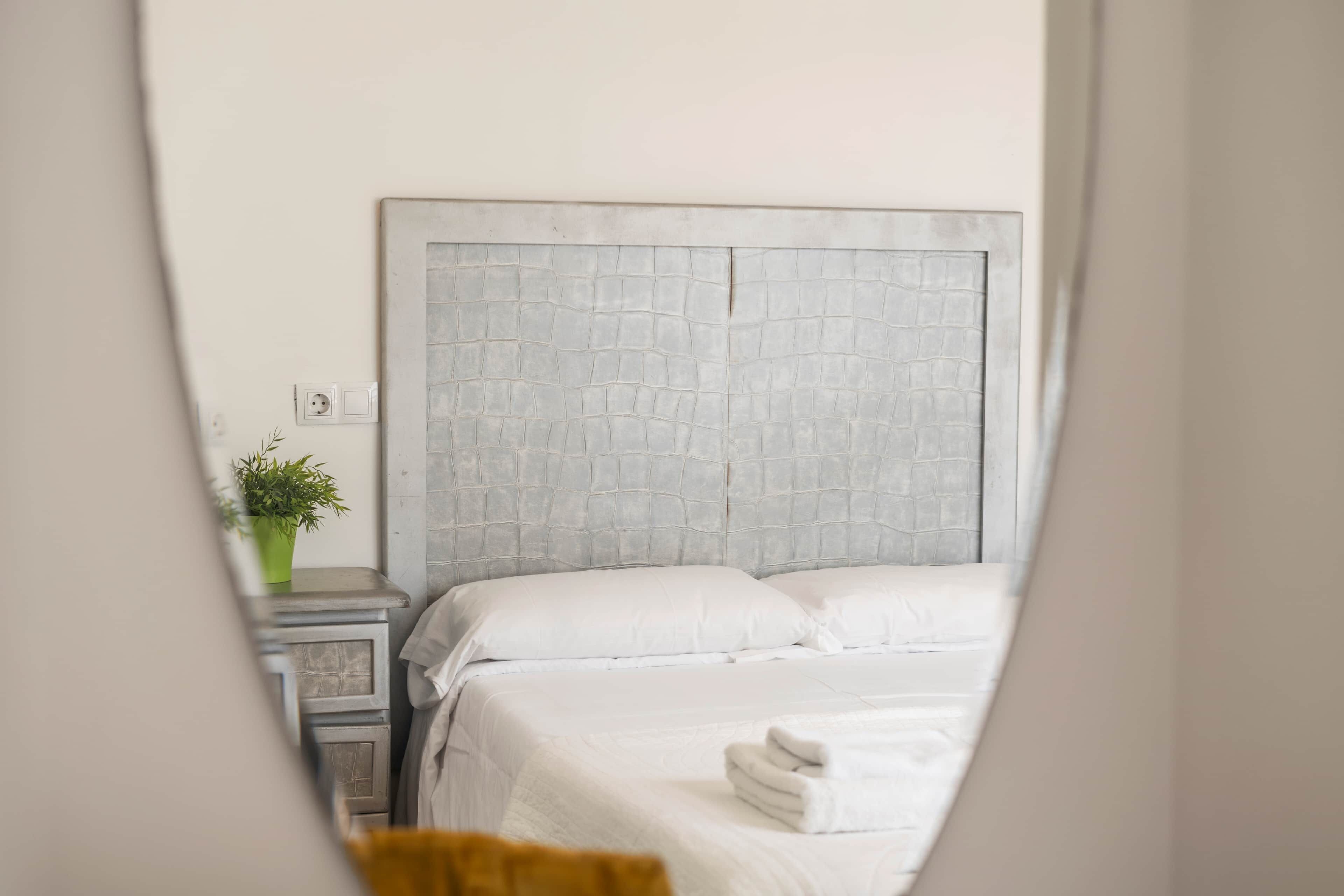 A bedroom with a headboard, a bedside table, a nightstand, a potted plant, and a bed with white bedding and a folded towel.