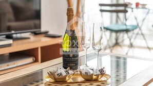 A bottle of champagne, two glasses, and chocolates on a table in a modern living room with a TV and a dining area in the background. - Ocean View Apartment (Platja de Gandia)
