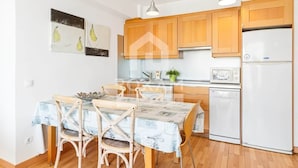 A modern kitchen with wooden cabinets, a white refrigerator, a microwave, and a dishwasher. There is a dining table with chairs and a potted plant on the counter. - Ocean View Apartment (Platja de Gandia)