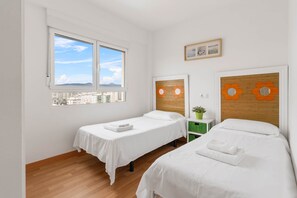 Quarto de hotel com duas camas de solteiro, cabeceira de madeira, janela com vista para a cidade e uma pequena planta verde em uma mesa de cabeceira.