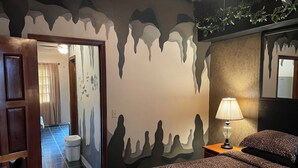 Ein Hotelzimmer mit Bett, Nachttisch mit Lampe, Badezimmer mit Toilette und Waschbecken sowie ein Flur mit einer Tür zu einem weiteren Zimmer.