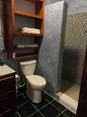 A bathroom with a toilet, a shelf with folded towels, a shower with mosaic tiles, and a dark tiled floor.